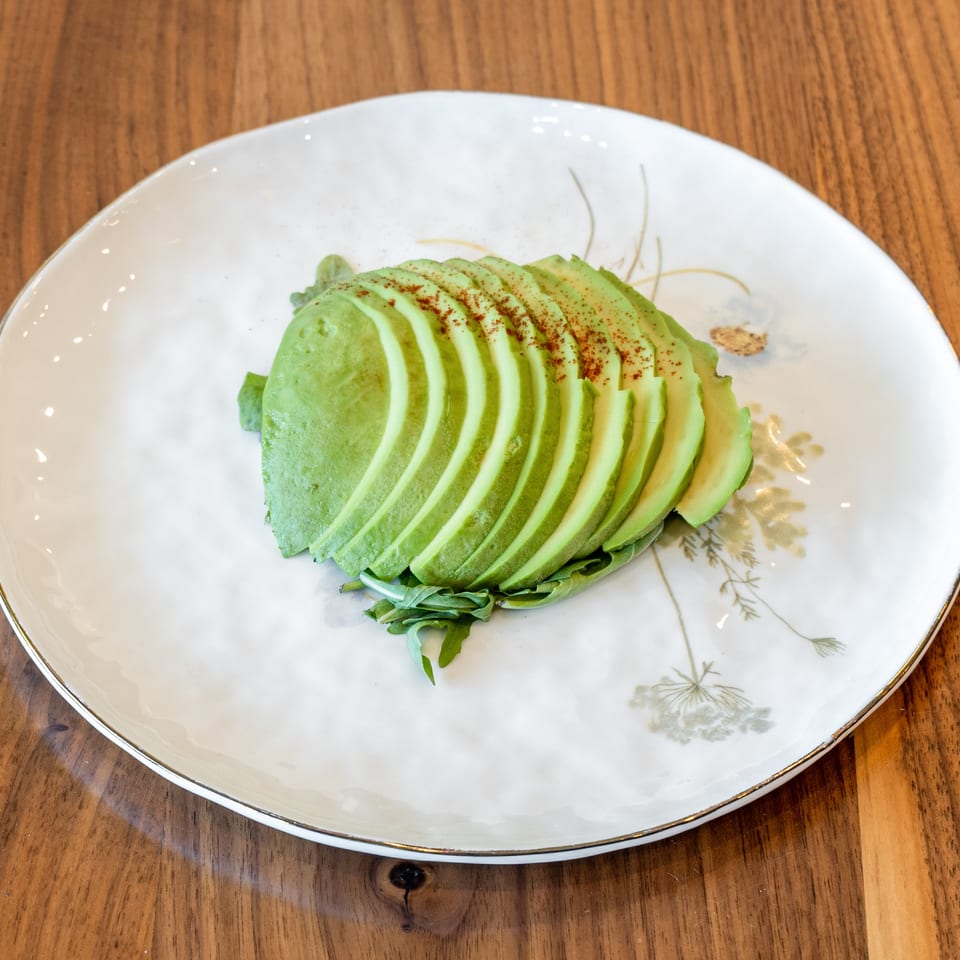Sliced avocado with paprika on a bed of arugula, served on a white plate with gold trim.