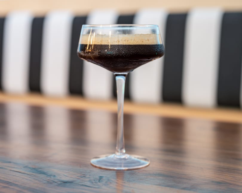 A dark, foamy espresso martini in a stemmed glass on a wooden table with a striped background.