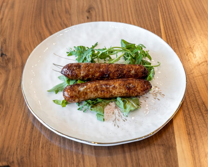 Two grilled sausages on a bed of fresh arugula, served on a white plate with gold trim.