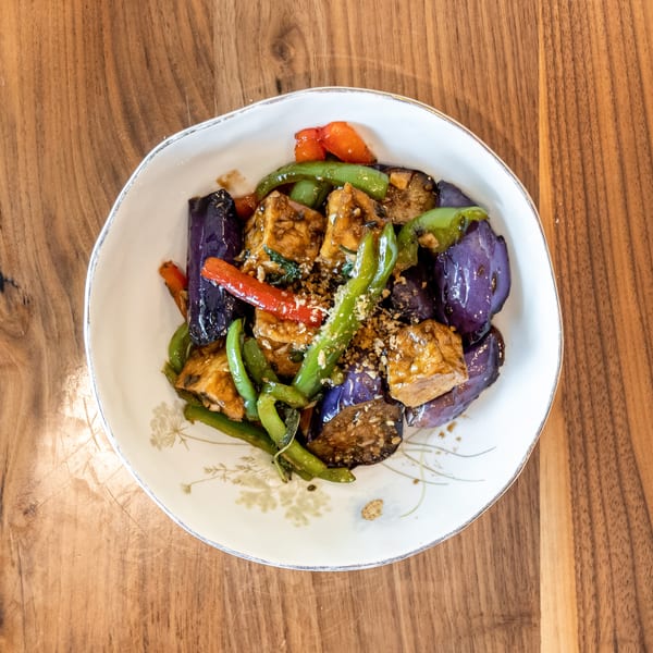 Stir-fried eggplant, green beans, red peppers, and tofu in a white bowl on a wooden table.