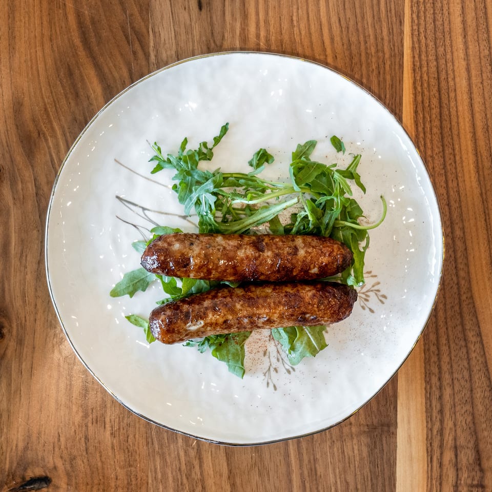 Two grilled sausages on a bed of fresh arugula, served on a white plate with gold trim.