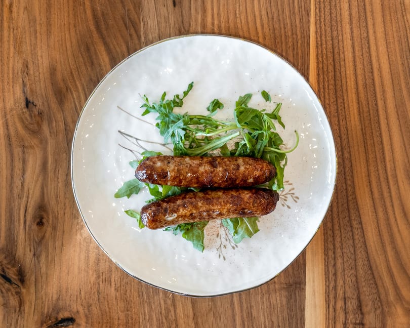 Two grilled sausages on a bed of fresh arugula, served on a white plate with gold trim.
