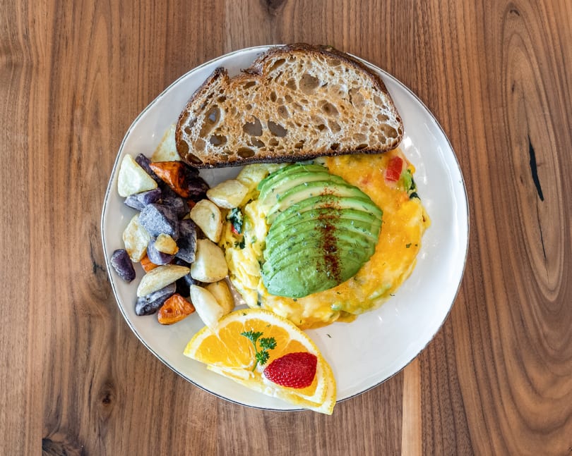 Breakfast plate with omelet topped with avocado, roasted potatoes, sourdough toast, and fruit.