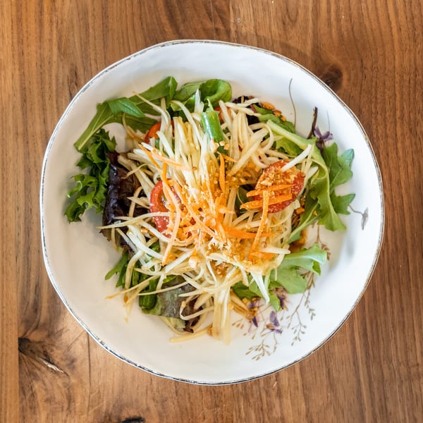 A vibrant papaya salad with shredded carrots, tomatoes, and greens in a white bowl on a wooden table.