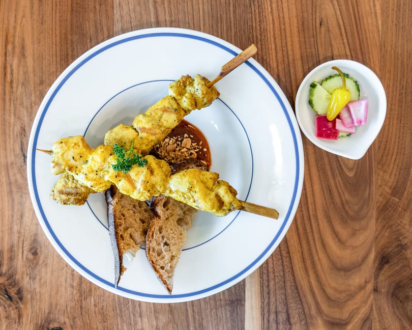 Two chicken satay skewers with peanut sauce, bread, and a side of pickled vegetables.