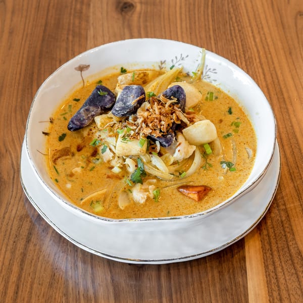 Creamy yellow curry soup with purple potatoes, fried onions, and fresh herbs in a white bowl.