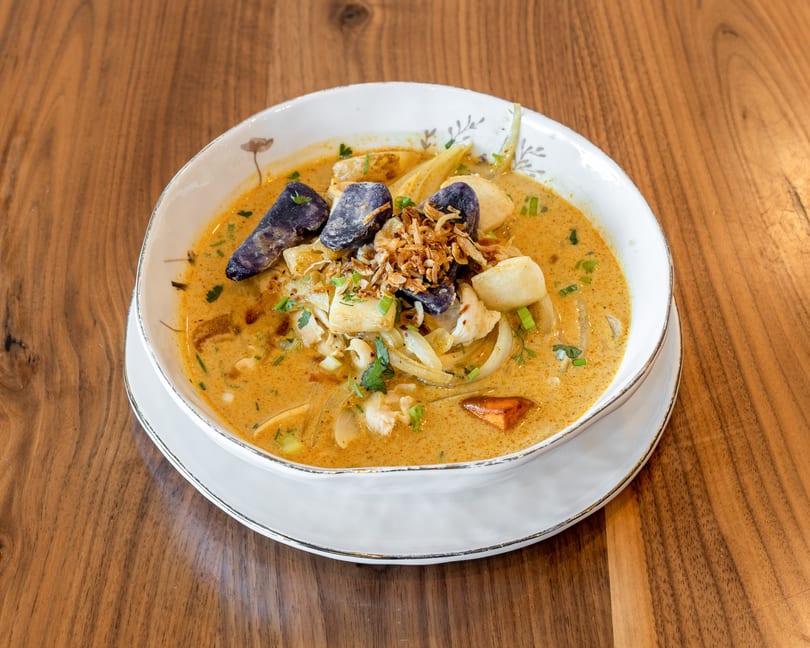 Creamy yellow curry soup with purple potatoes, fried onions, and fresh herbs in a white bowl.