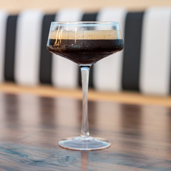 A dark, foamy espresso martini in a stemmed glass on a wooden table with a striped background.