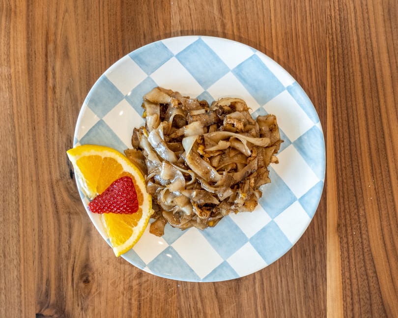 Noodle dish with a side of orange and strawberry on a blue and white checkered plate.