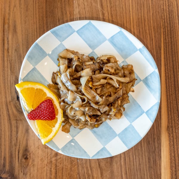 Noodle dish on a blue and white checkered plate, garnished with an orange slice and strawberry.