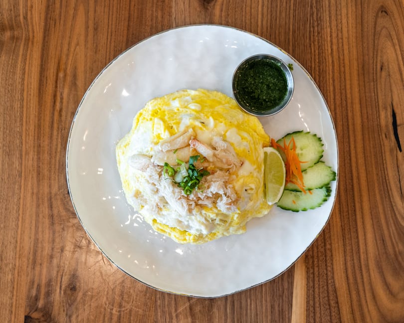 Crab omelet with green sauce, lime, and cucumber slices on a white plate, on a wooden table.