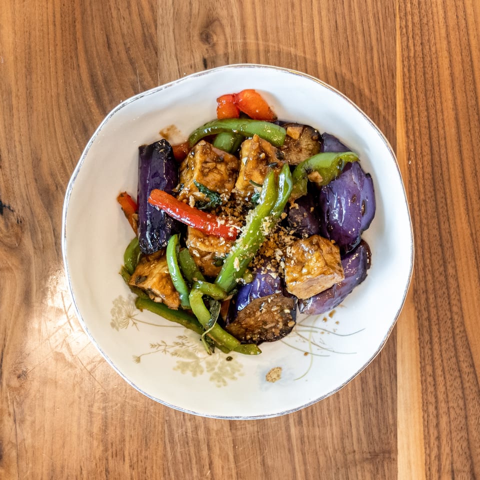 Stir-fried eggplant, green beans, red peppers, and tofu in a white bowl on a wooden table.