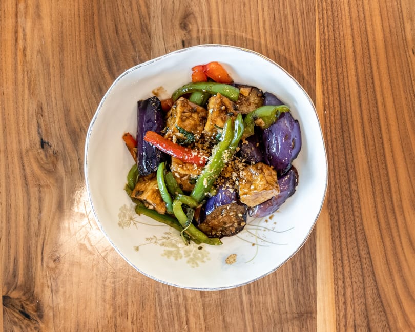 Stir-fried eggplant, green beans, red peppers, and tofu in a white bowl on a wooden table.
