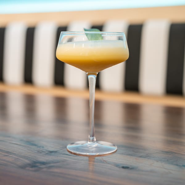 Yellow cocktail with a green leaf garnish on a wooden table with striped seating.
