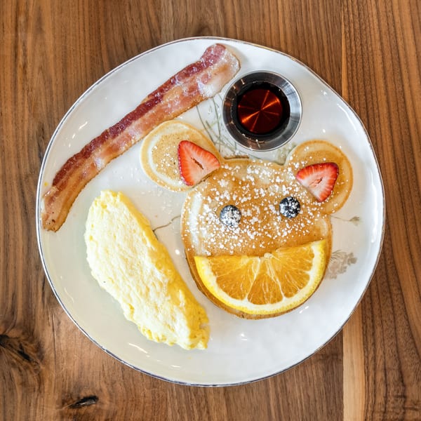 A bear-shaped pancake with fruit, bacon, scrambled eggs, and syrup on a white plate.