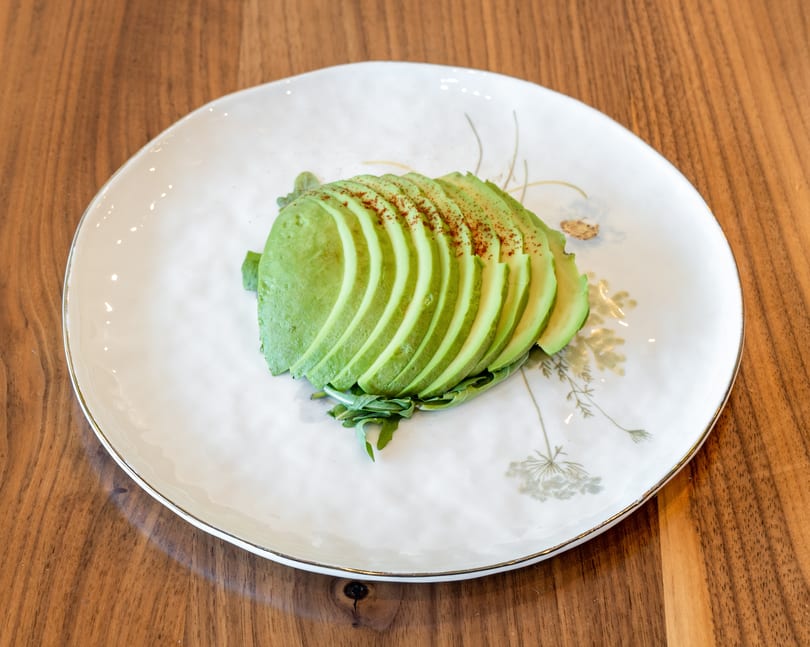 Sliced avocado with paprika and greens on a white plate with gold rim, on a wooden table.