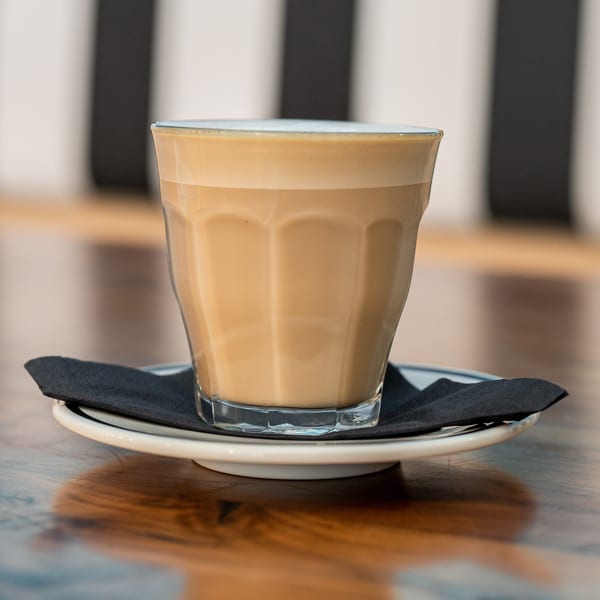A latte in a clear glass with a black napkin on a white saucer, on a wooden table.