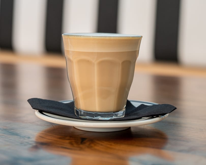 A latte in a clear glass with a black napkin on a white saucer, on a wooden table.
