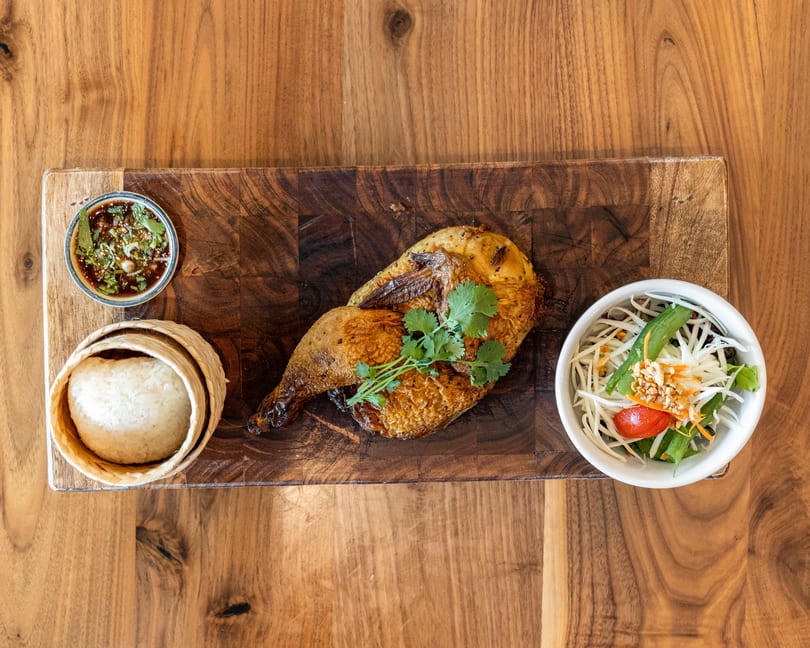 Grilled chicken, sticky rice, papaya salad, and dipping sauce on a wooden board.