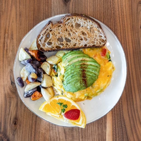Breakfast plate with omelet topped with avocado, roasted potatoes, sourdough toast, and fruit.