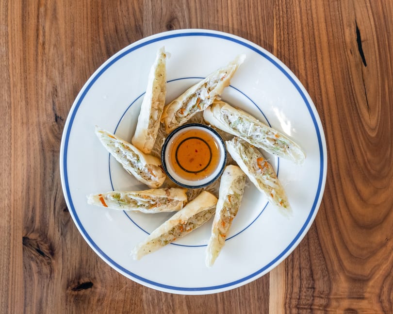 Crispy spring rolls with vegetable filling and a side of dipping sauce on a white plate.