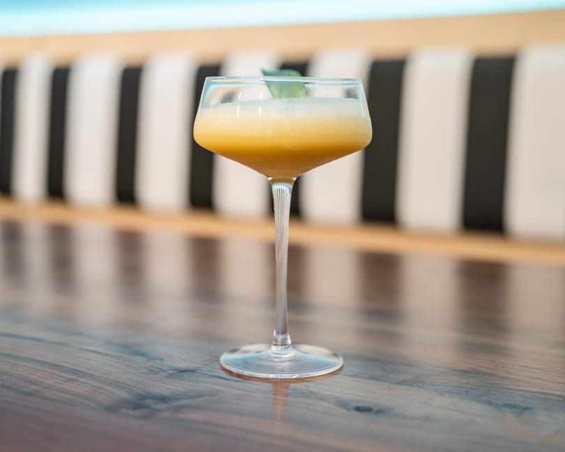 Yellow cocktail with a green leaf garnish on a wooden table with striped seating.