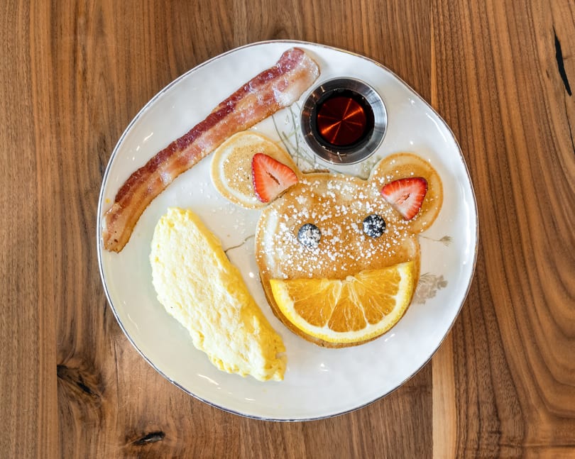 A bear-shaped pancake with fruit, bacon, scrambled eggs, and syrup on a white plate.