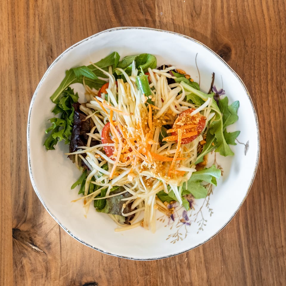A vibrant papaya salad with shredded carrots, tomatoes, and greens in a white bowl on a wooden table.
