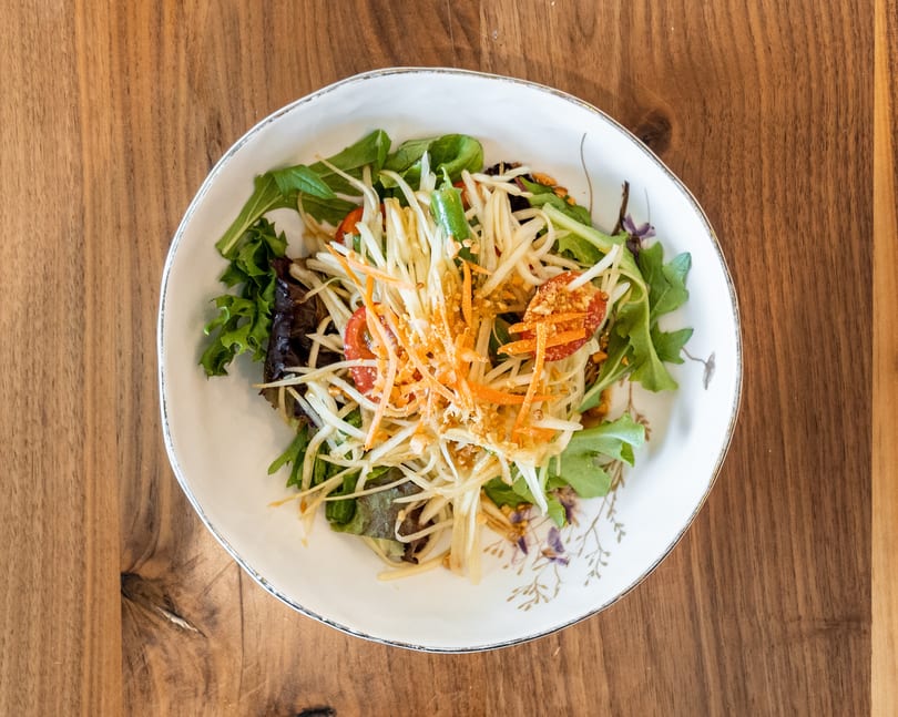 A vibrant papaya salad with shredded carrots, tomatoes, and greens in a white bowl on a wooden table.
